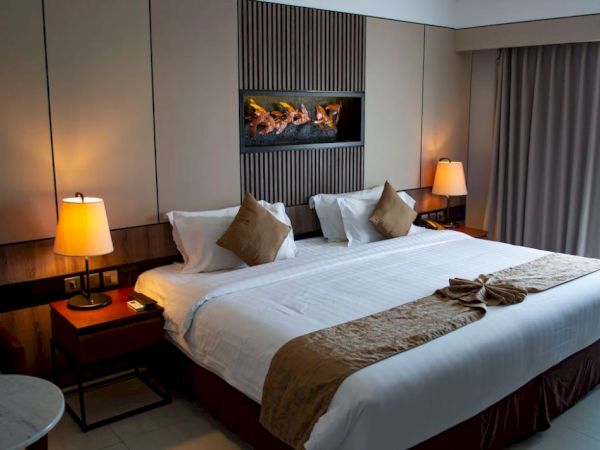 A well-lit hotel room with a large bed, two nightstands with lamps, decorative pillows, wall decor, and a window with curtains is shown.