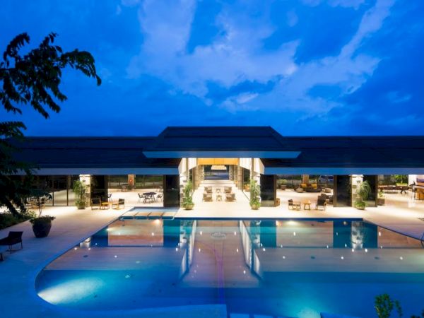 An illuminated outdoor pool with lounge seating and a modern building in the background, surrounded by trees, with a blue evening sky.