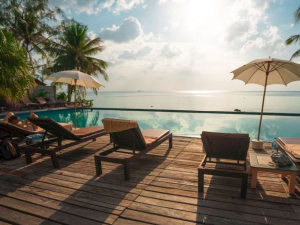 Poolside scene with lounge chairs, umbrellas, and palm trees overlooking a calm ocean at sunrise or sunset, under a partly cloudy sky.