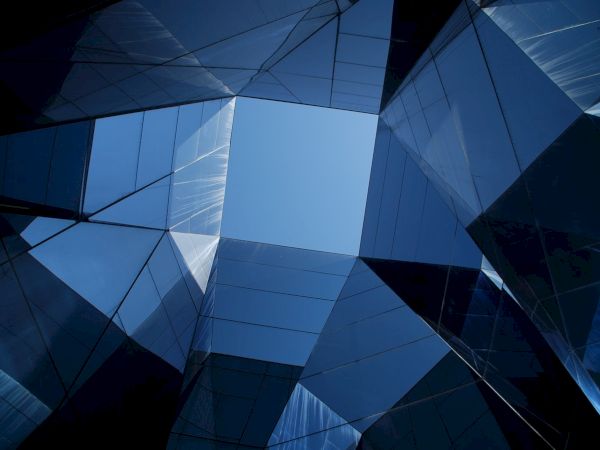The image shows an upward view of modern glass buildings with geometric shapes, reflecting the blue sky in the center of the composition.