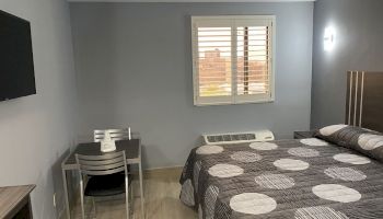 The image shows a minimalist hotel room with a bed, a small table and two chairs, a wall-mounted TV, and a window with shutters.