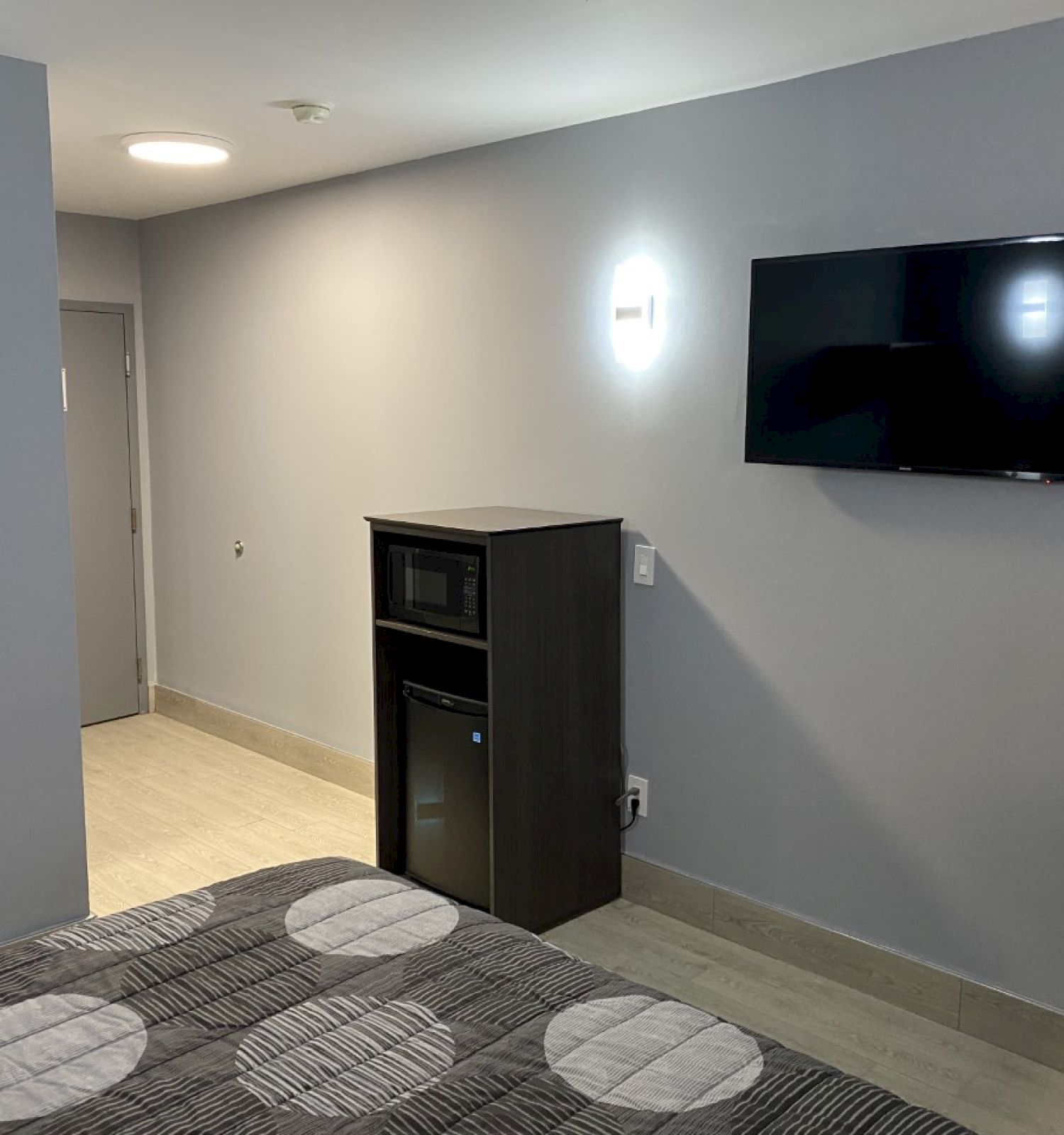 The image shows a modern, minimalistic hotel room with a bed, wall-mounted TV, small fridge, microwave, desk, and chair in a light-colored setting.