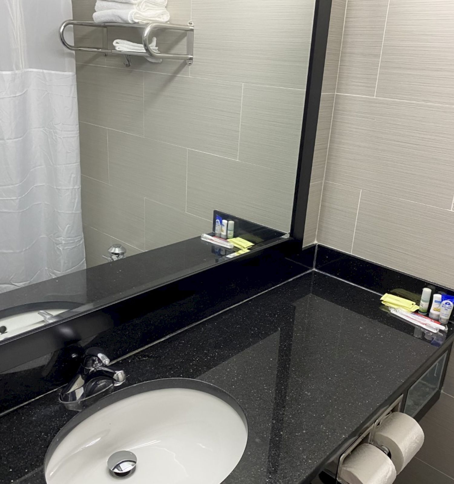 A clean bathroom with a black countertop, sink, large mirror, toiletries, a towel on a rack, and a shower with a white curtain.