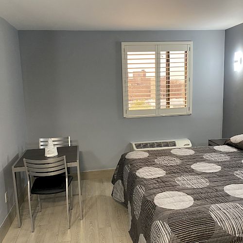 A modern, minimalist bedroom with a bed, desk and chairs, wall-mounted TV, and window with blinds, all in a neutral color palette.