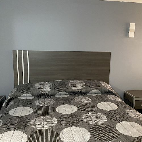 The image shows a bedroom with a bed covered in a gray patterned bedspread. Two bedside tables and wall-mounted lights are visible.