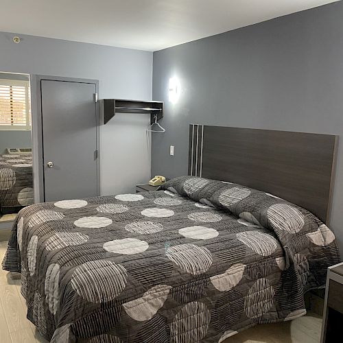 A modern, minimalistic hotel room with a double bed, gray patterned bedding, a nightstand, and a mirror reflecting the space, creating a clean vibe.