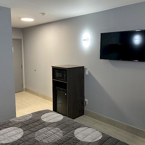 A hotel room with a bed, wall-mounted TV, dresser with microwave and mini-fridge, small table, chair, and modern lighting is shown in the image.