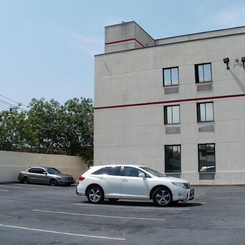 A nearly empty parking lot with two parked cars, one white and one gray, adjacent to a beige building with windows and security cameras.