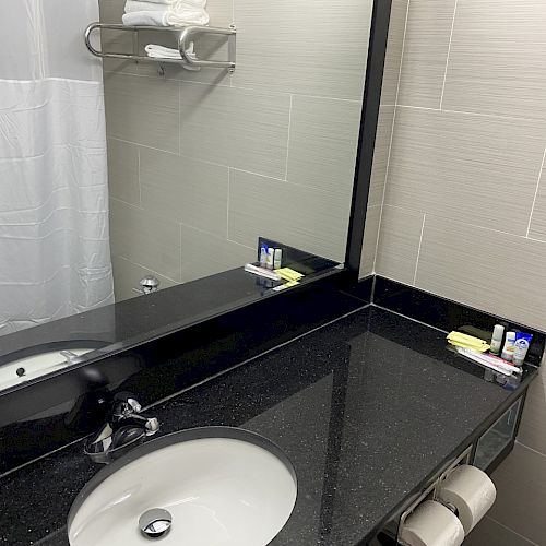 A bathroom with a large mirror, countertop sink, black vanity, towel rack with folded towels, and various toiletries placed around the counter.