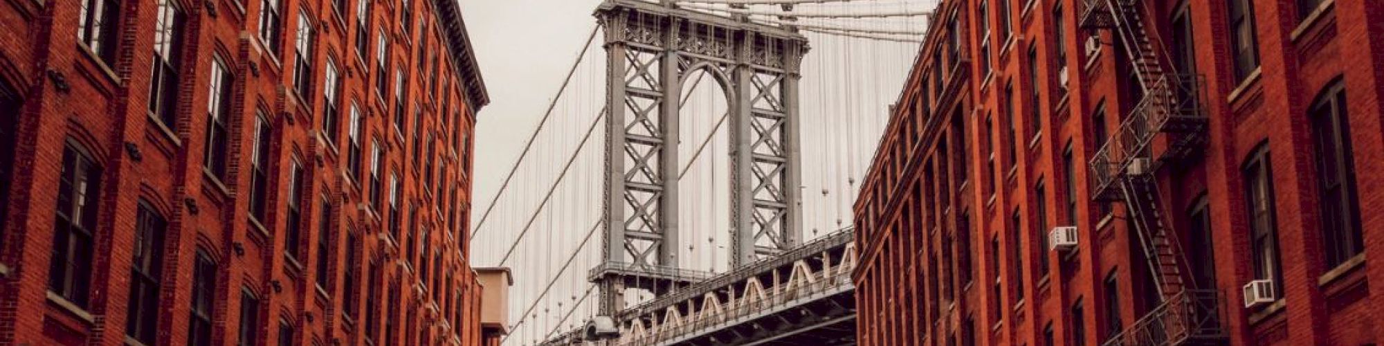 A view of a suspension bridge framed by red brick buildings on both sides, with street signs visible at the intersection.