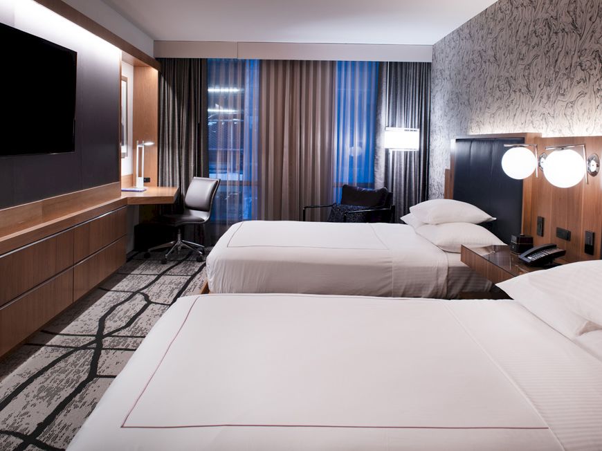 A modern hotel room with two large beds, a wall-mounted TV, a desk with a chair, and contemporary decor, featuring a large window with curtains.