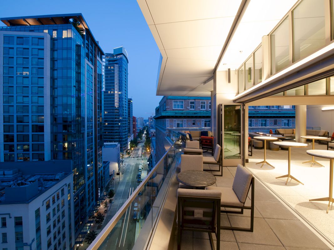 An outdoor terrace with seating overlooks a cityscape of tall buildings at dusk. Large windows provide a view into the well-lit interior.