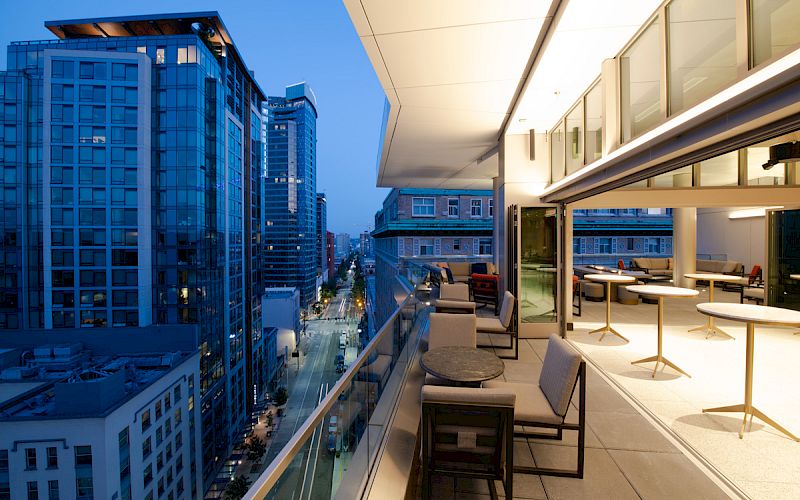 A modern balcony with chairs and tables overlooks a cityscape filled with tall buildings at dusk, while the interior is warmly lit.